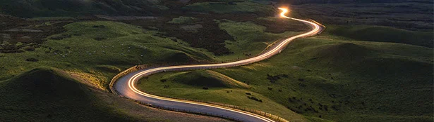 Winding road cutting through rolling green hills at dusk, with long-exposure light trails from passing vehicles creating a glowing path across the landscape.