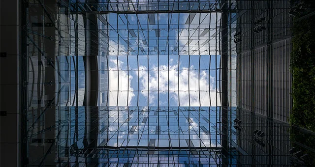 Upward view from inside a modern glass atrium, showing a grid of steel and glass panels framing a bright sky with clouds above, surrounded by tall building walls and reflective surfaces.
